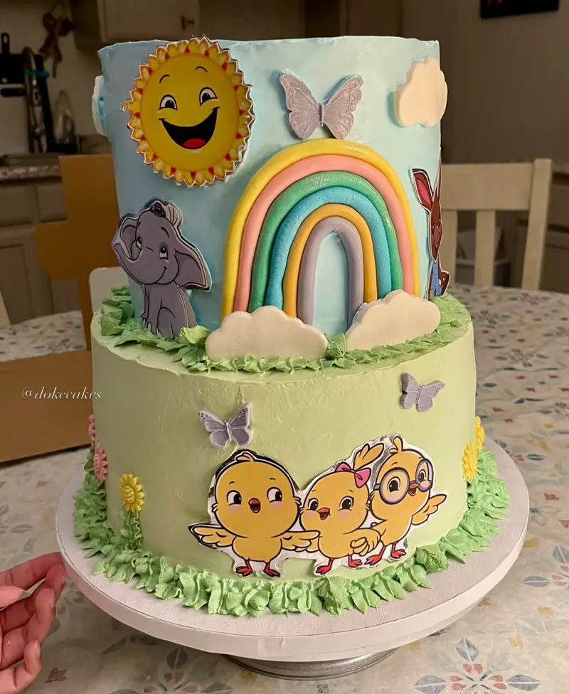 Two-tier pastel cake with a rainbow, sunshine, an elephant and baby chick characters