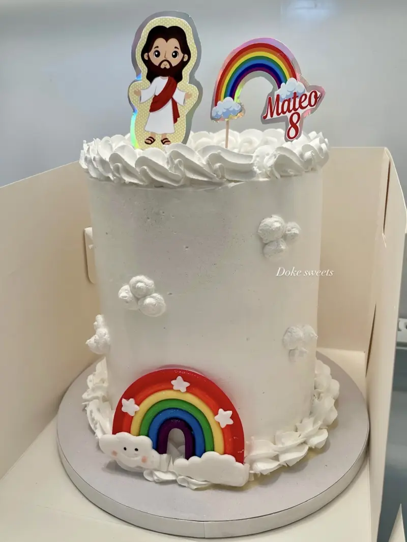 Tall white cake with rainbows, clouds and a Jesus topper